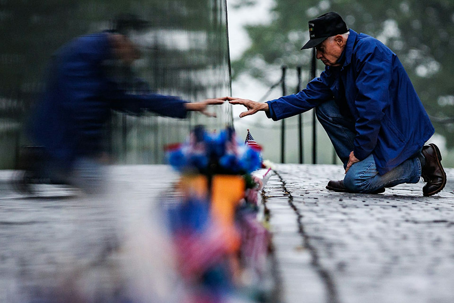 A kneeling elderly man wearing a blue jacket and a baseball cap extends his arm to touch a memorial.