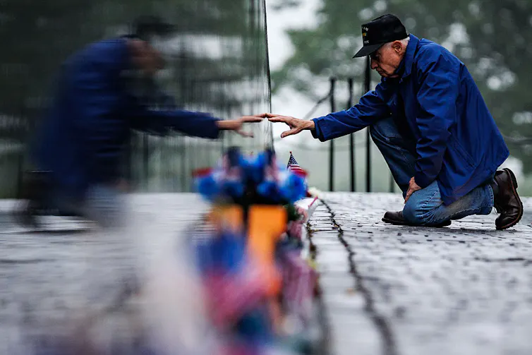 A kneeling elderly man wearing a blue jacket and a baseball cap extends his arm to touch a memorial.