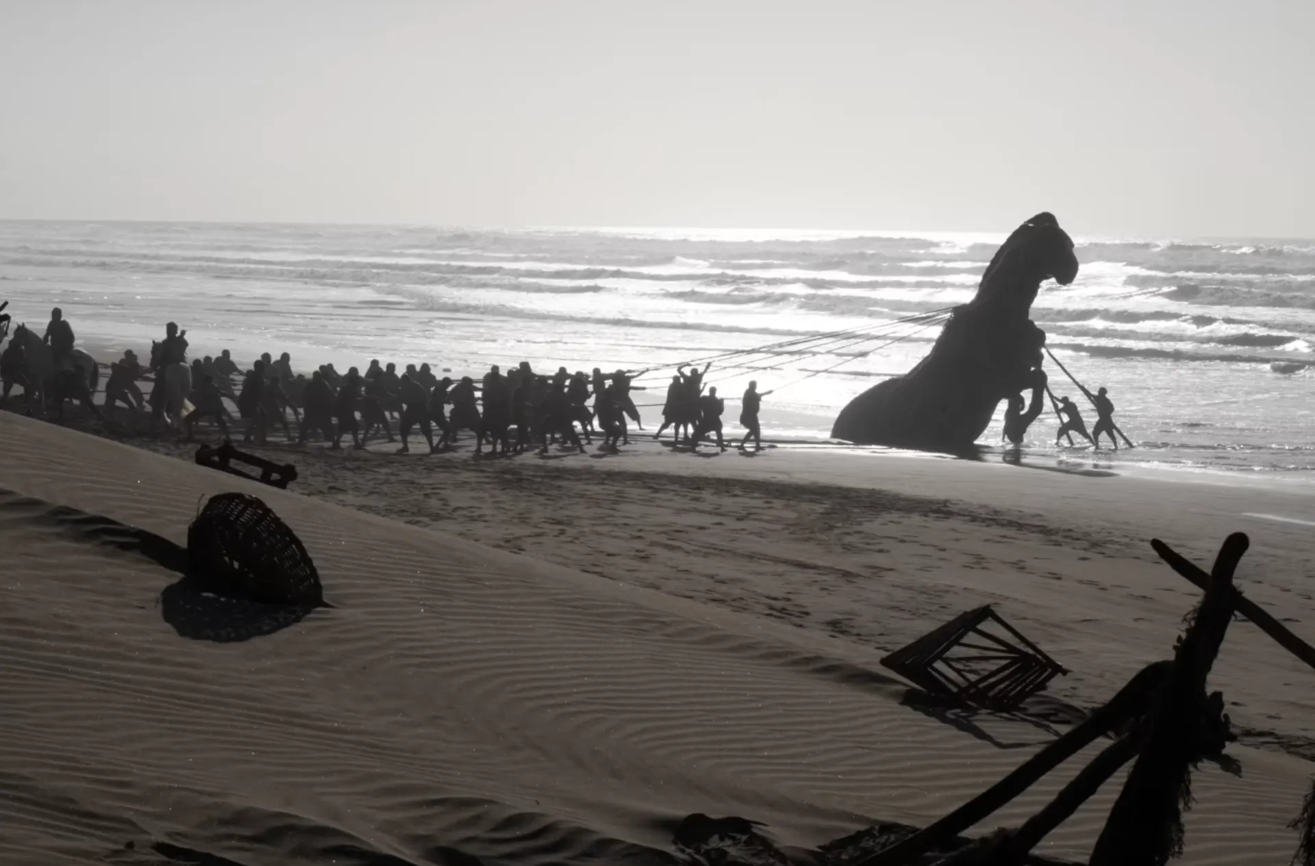 Silhouette of a large group of men pulling a massive wooden horse that appears to have washed up on the beach.