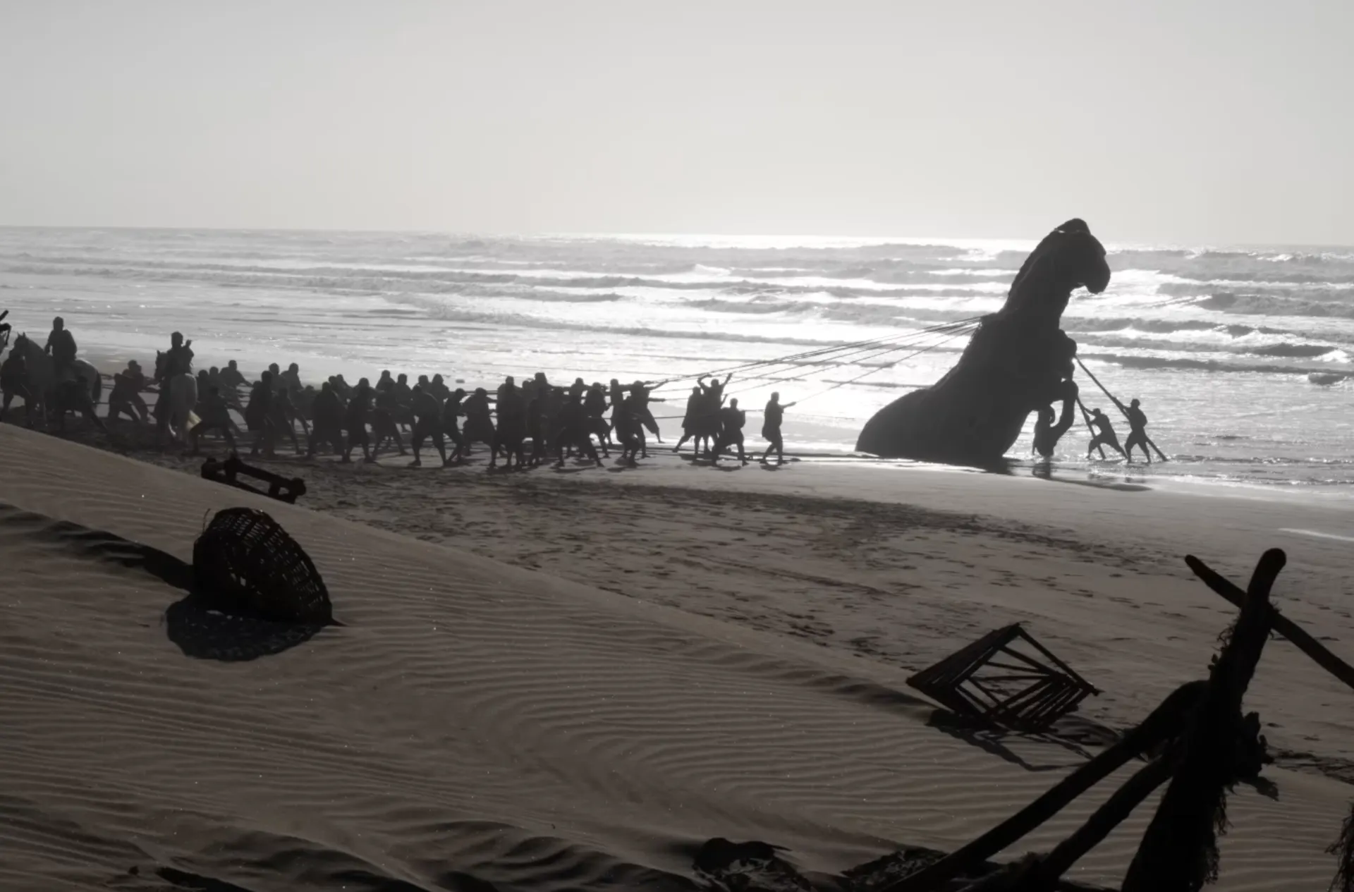 Silhouette of a large group of men pulling a massive wooden horse that appears to have washed up on the beach.