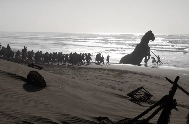 Silhouette of a large group of men pulling a massive wooden horse that appears to have washed up on the beach.