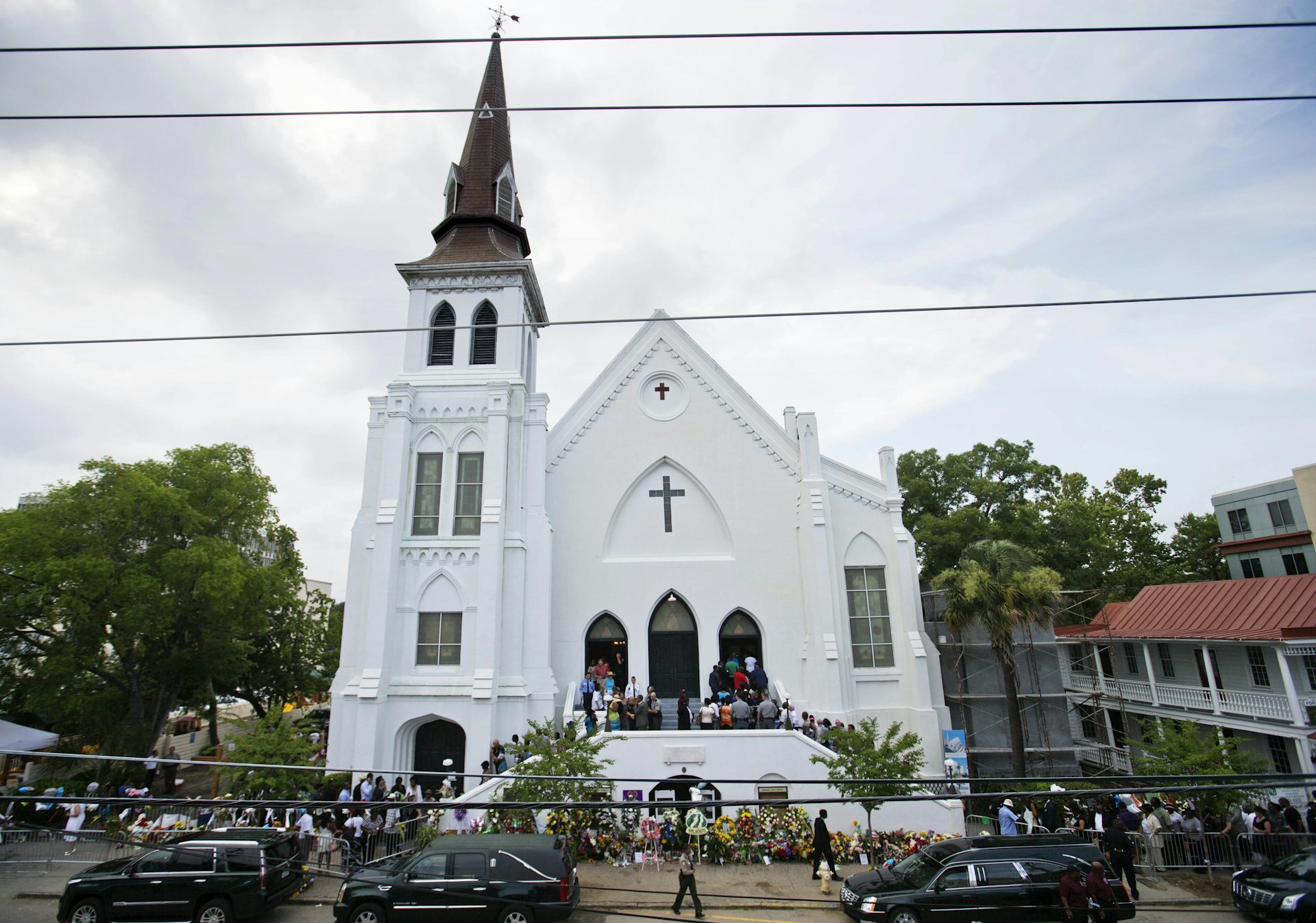 A white church with dozens of people lined up outside, waiting to enter. A memorial with flowers lines the sidewalk.