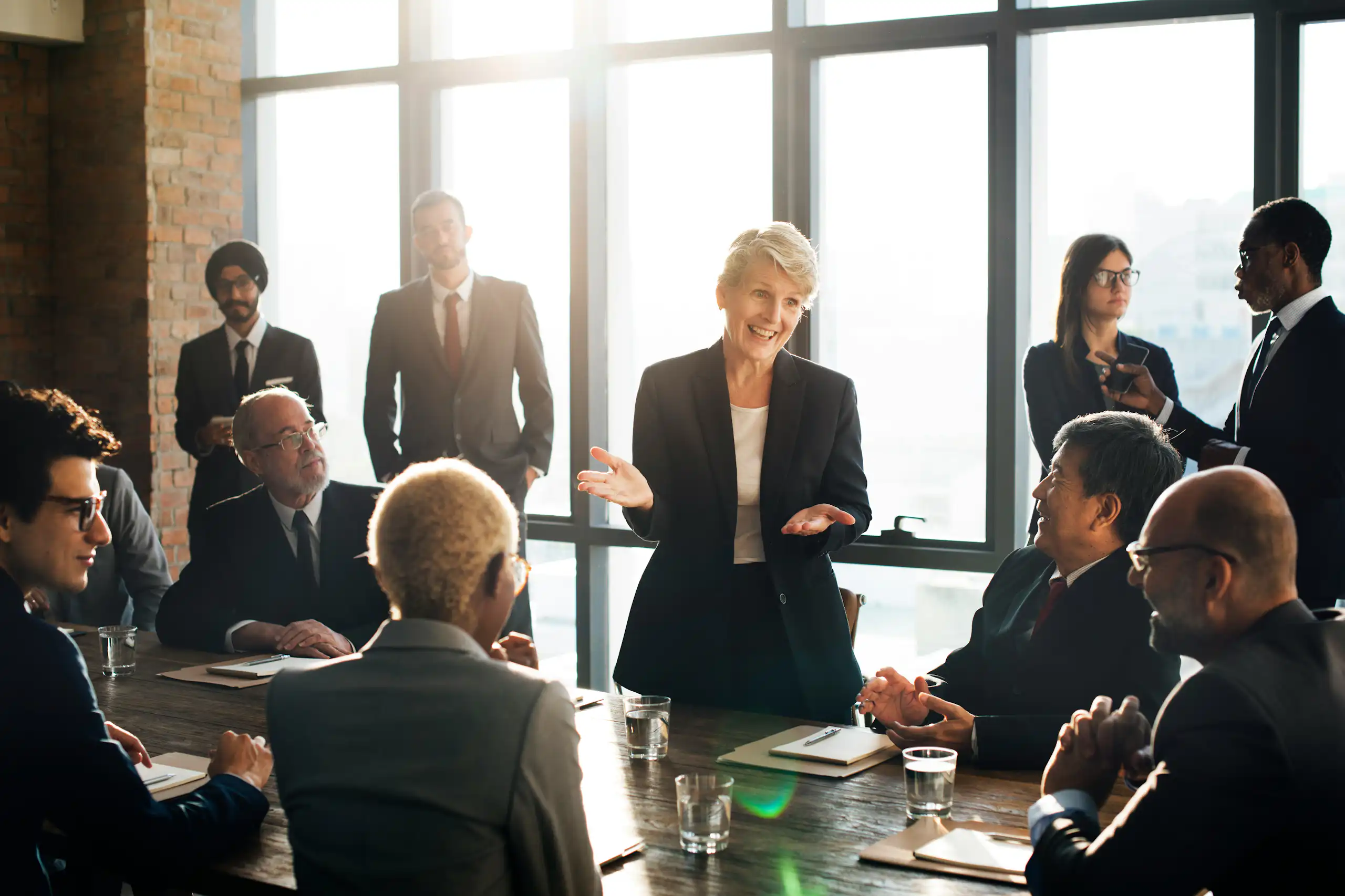 A woman standing is speaking to the other participants in a meeting—men and women—some of whom are seated at a conference table, while others are standing next to her