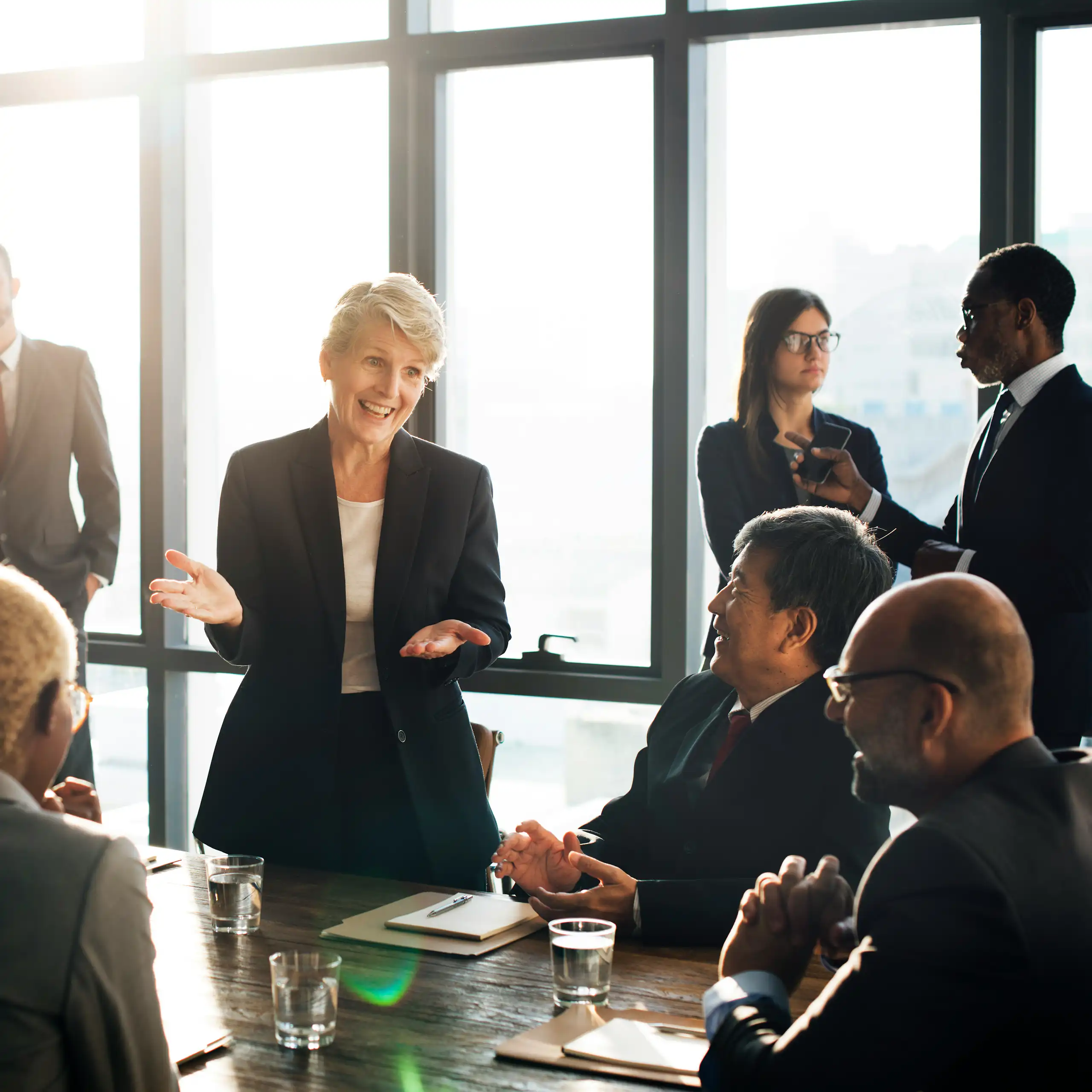A woman standing is speaking to the other participants in a meeting—men and women—some of whom are seated at a conference table, while others are standing next to her