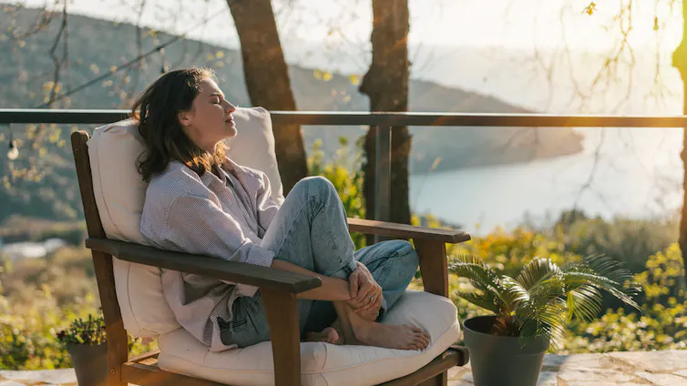 Mujer con los ojos cerrados sentada en una terraza con un paisaje costero al fondo