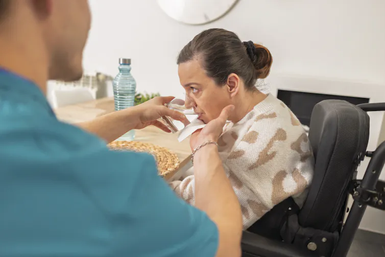 a carer helps a disabled woman to drink water from a glass.