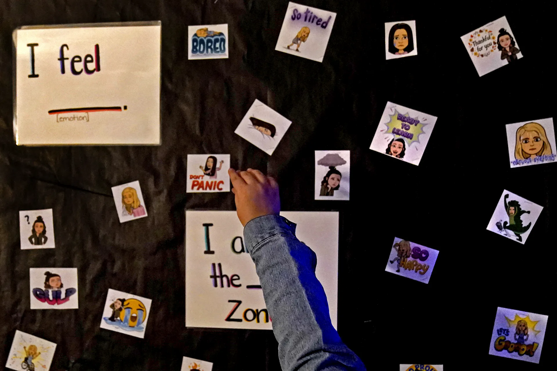 A young person's hand is seen touching a cut-out paper on a wall that says 'Don't panic.' There are other squares with emotions also on the wall, along with a sign that says 'I feel.'