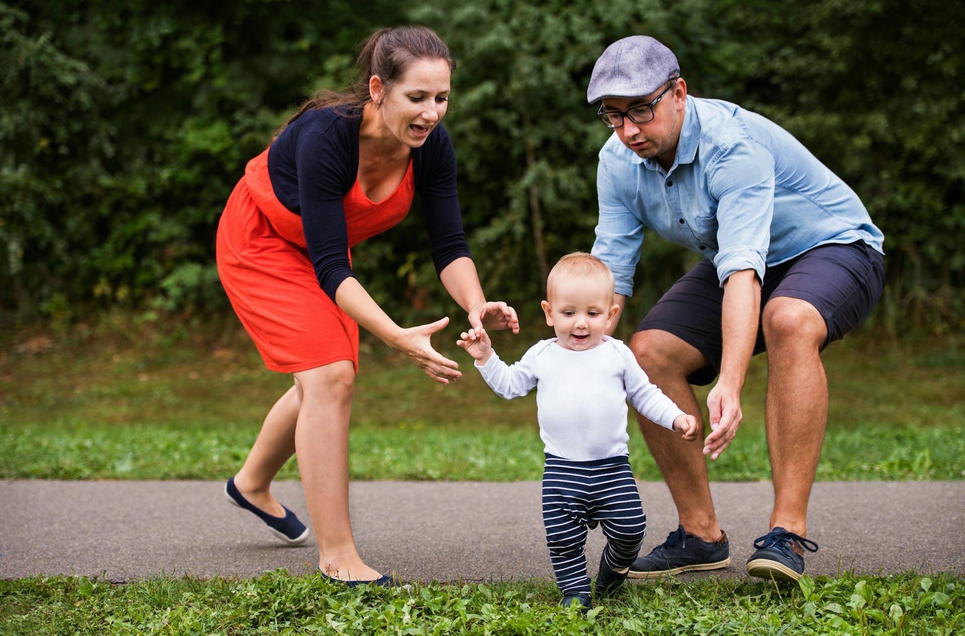 A couple are walking carefully besides a toddler who is learning to walk independently.