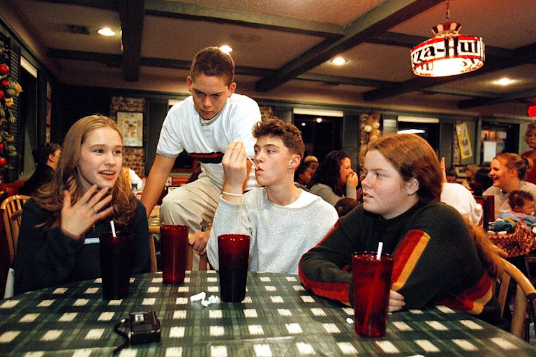 A vintage photo of a group of teenagers sits around a table in a Pizza Hut restaurant.