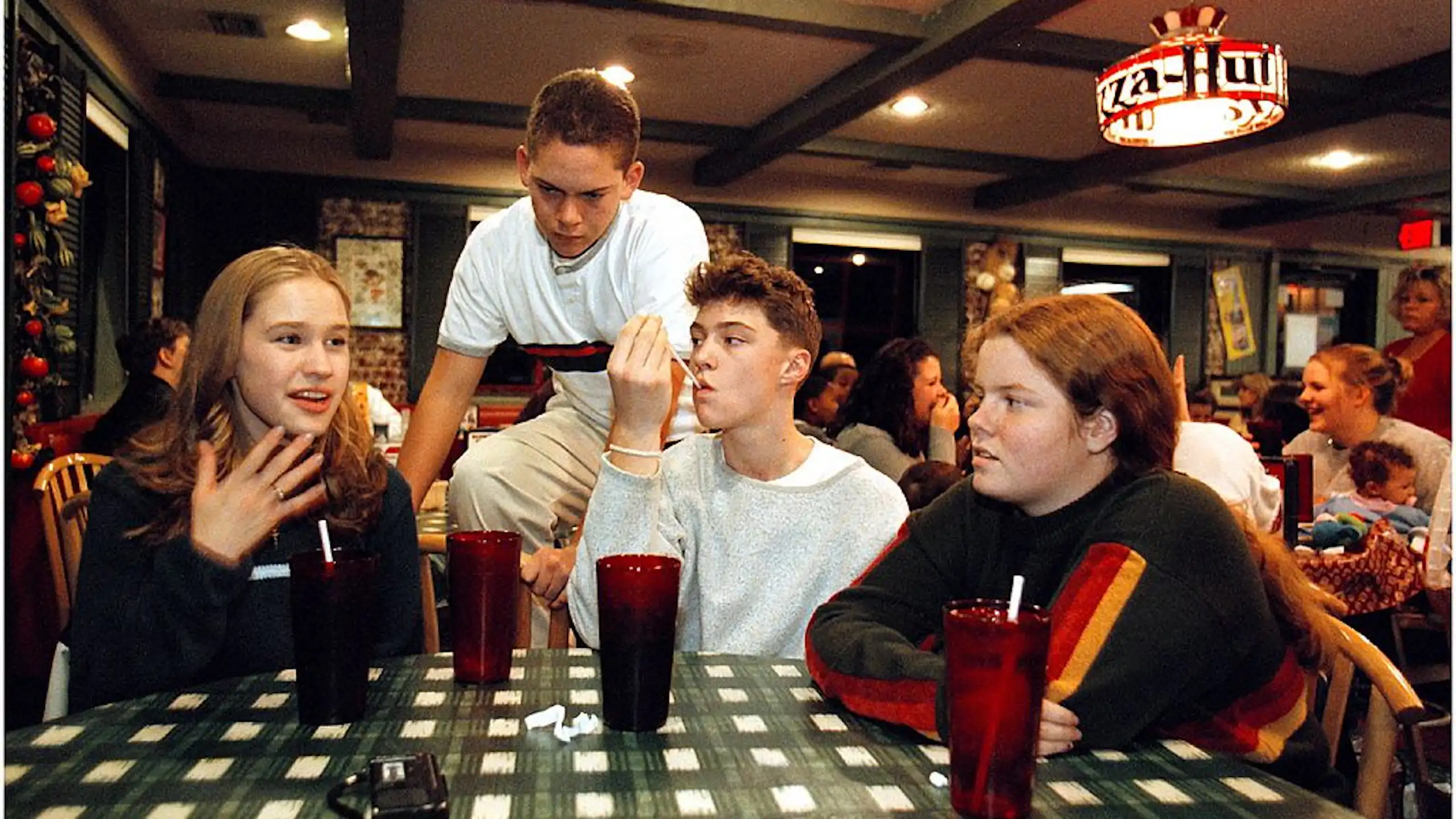 A vintage photo of a group of teenagers sits around a table in a Pizza Hut restaurant.