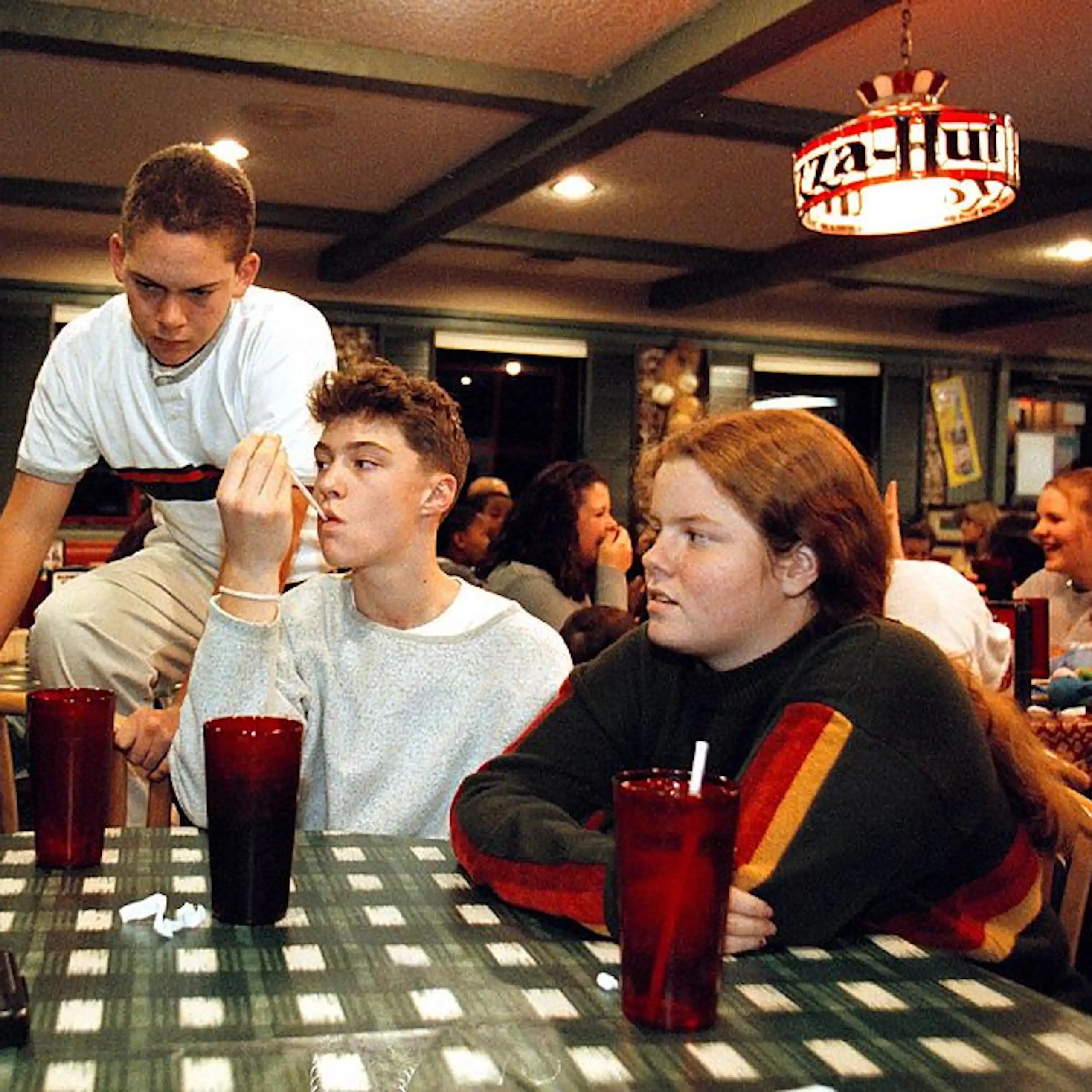 A vintage photo of a group of teenagers sits around a table in a Pizza Hut restaurant.