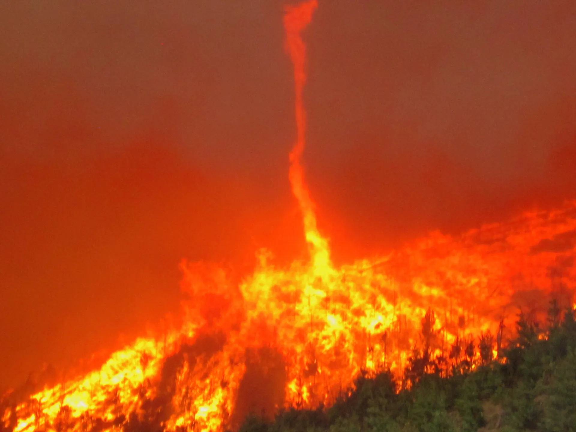 What looks like a thin tornado of fire rising from a wildfire in the forest below.