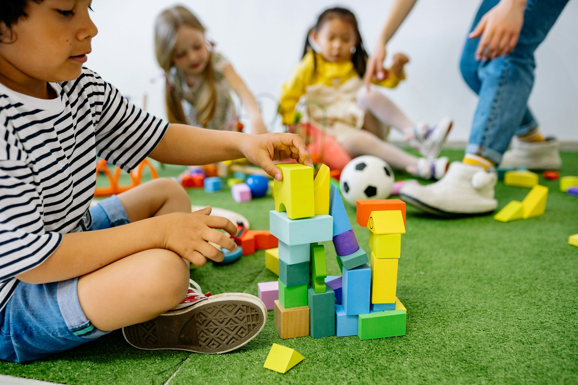 A child stacks blocks on a carpet with other children and an educator seen in soft focus in the background.