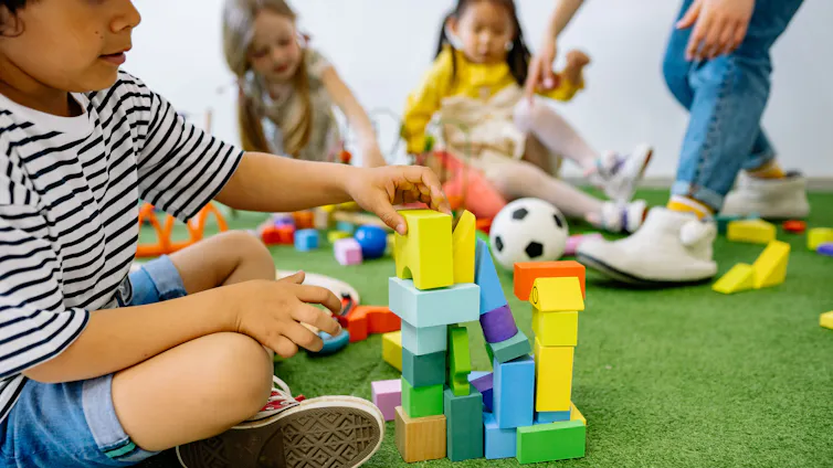 A child stacks blocks on a carpet with other children and an educator seen in soft focus in the background.