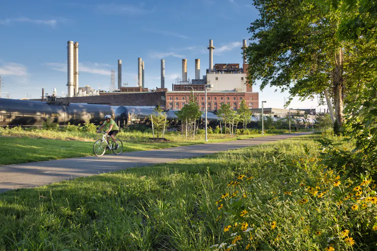 A view of cyclist on a trail surrounded by greenery with a factory in background