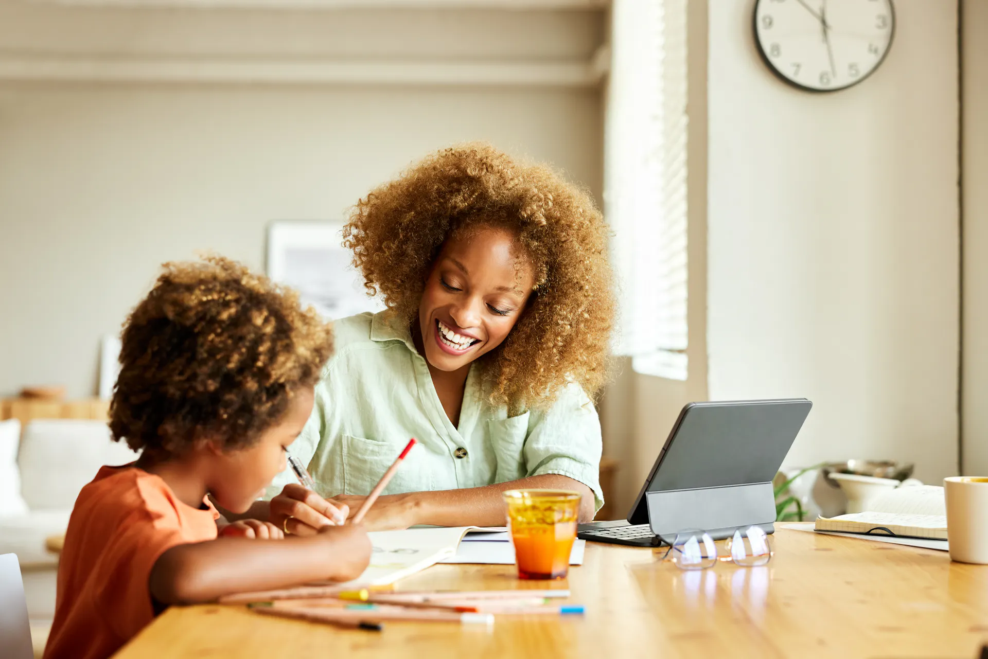 A smiling woman helps a young child draw at a dining table beside a tablet, notebooks, colored pencils, and a glass of juice in a bright home.