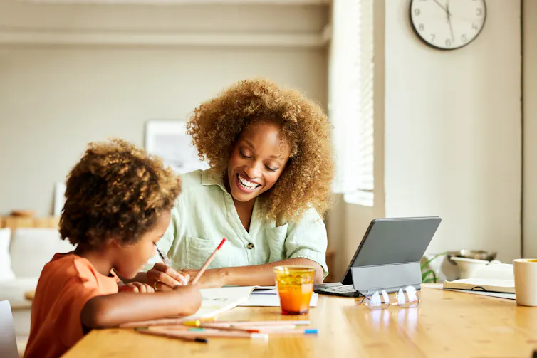 A smiling woman helps a young child draw at a dining table beside a tablet, notebooks, colored pencils, and a glass of juice in a bright home.