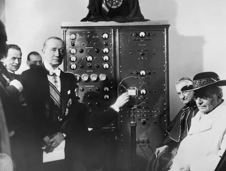 Black-and-white historical photograph of a formally dressed man, Guglielmo Marconi, demonstrating a radio microphone before a large control panel, while a seated elderly man in white robes and several onlookers watch.