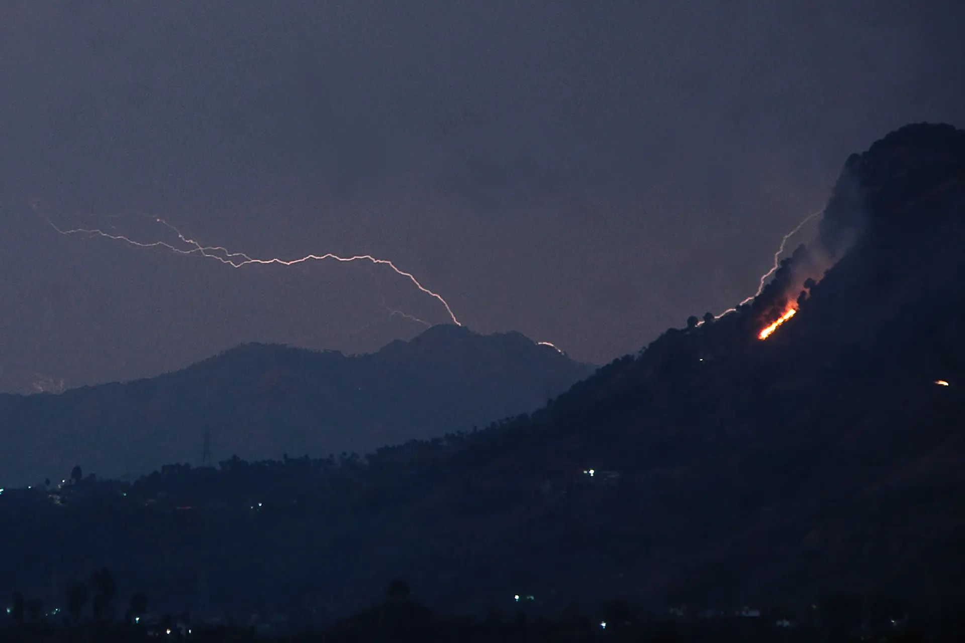 Lightning strikes behind a mountain as smoke bellows from a wildfire in the foreground.