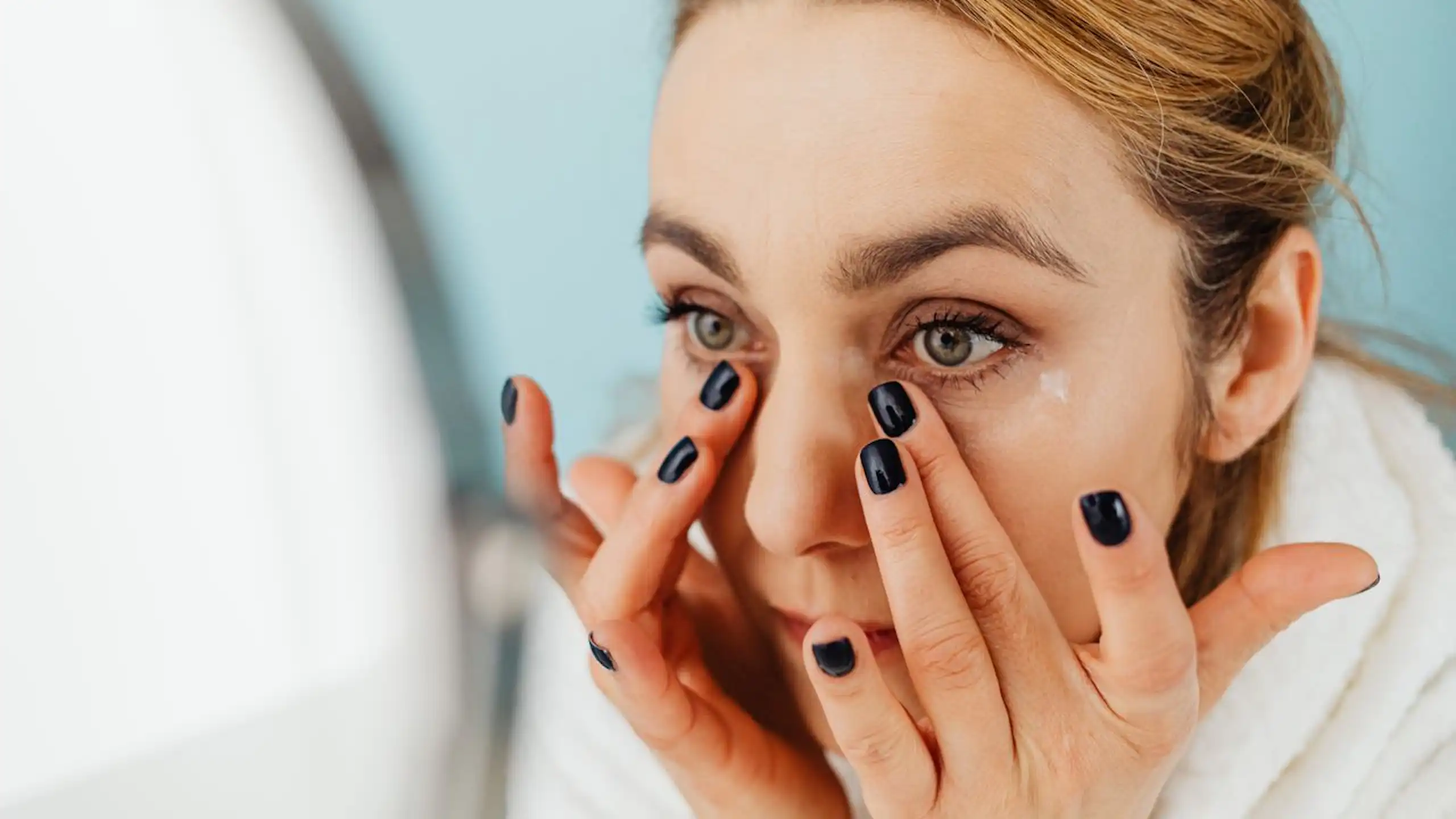 Woman applying skincare under eyes, looking in mirror