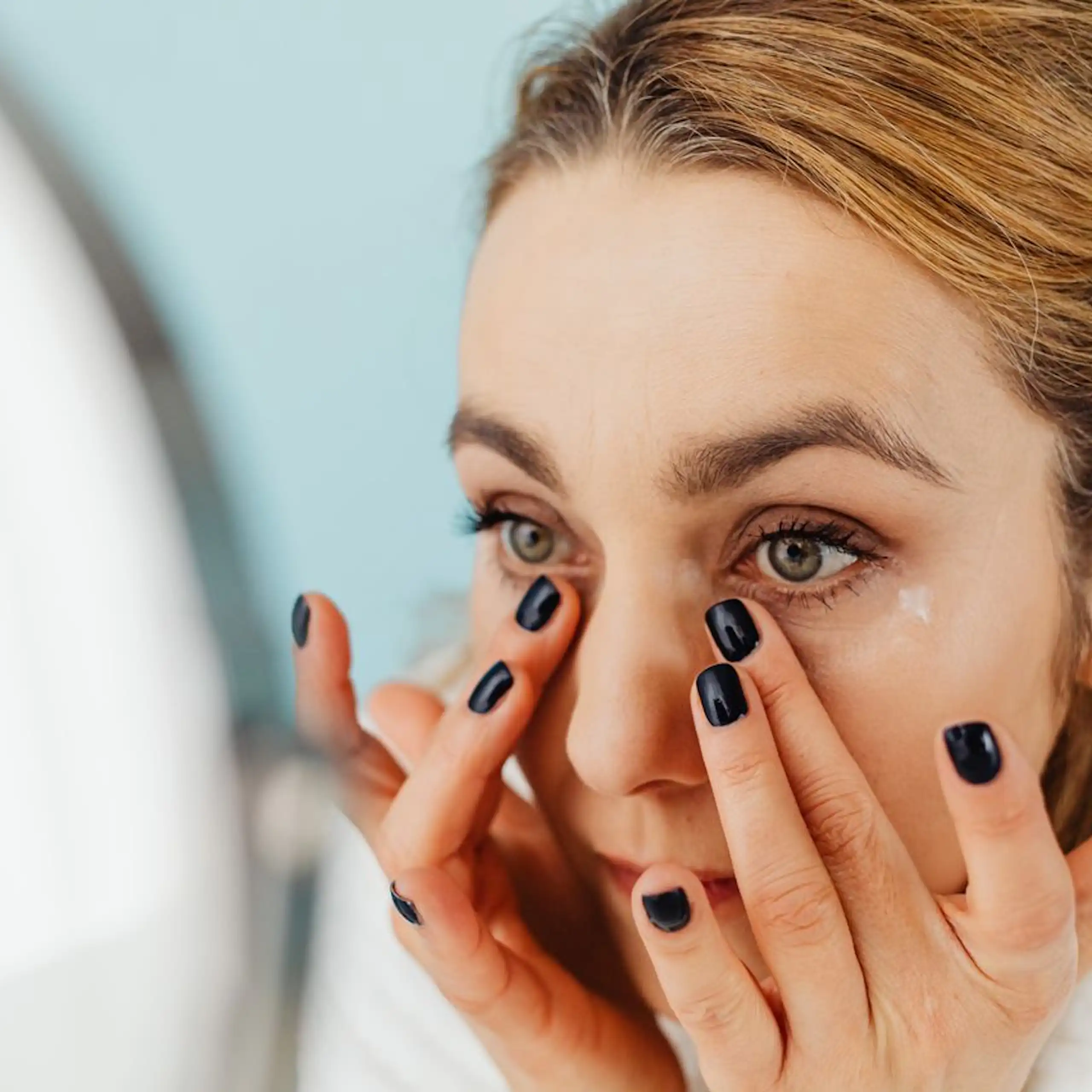 Woman applying skincare under eyes, looking in mirror