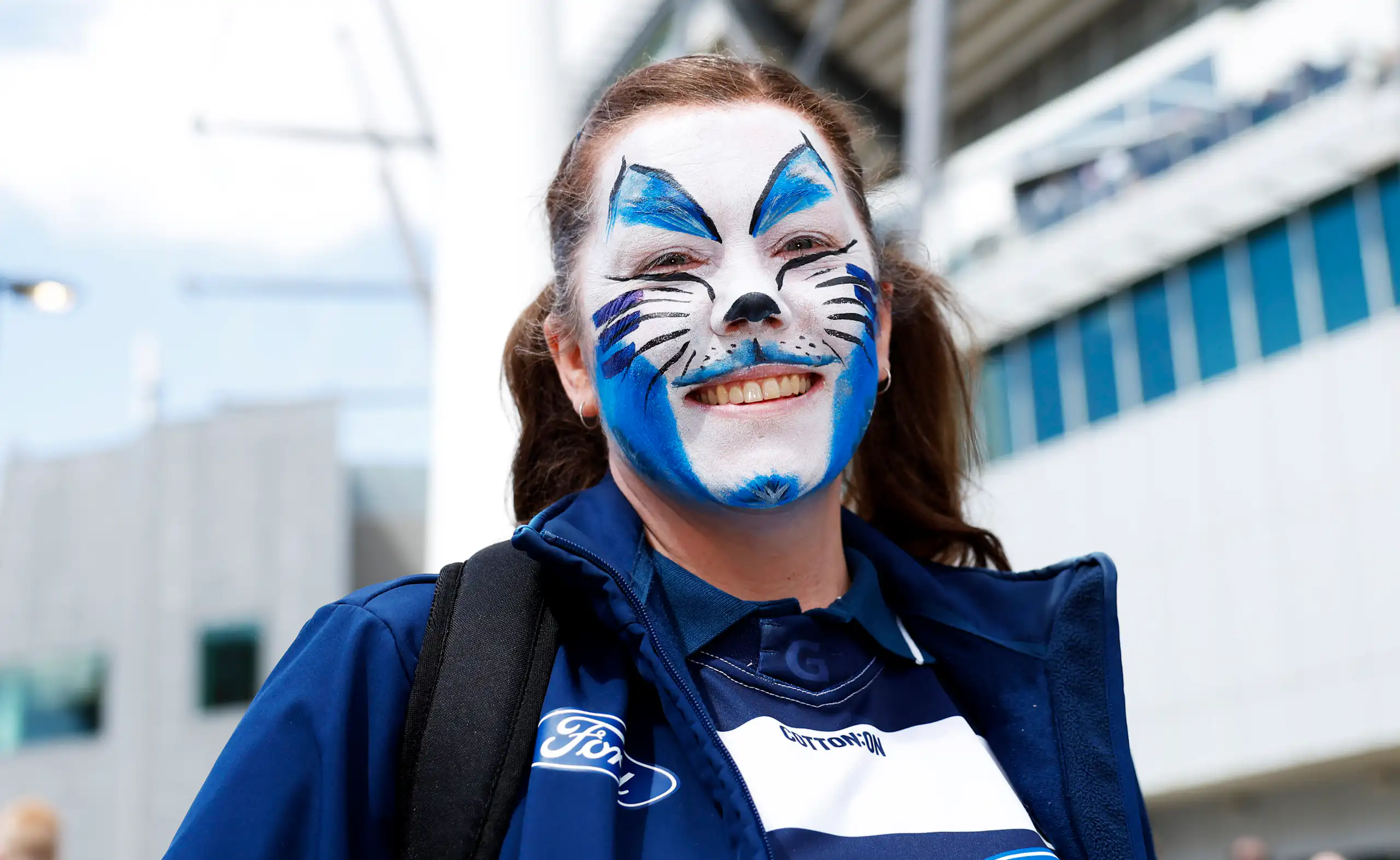 A young woman with blue and white cat face paint smiling outside a stadium.