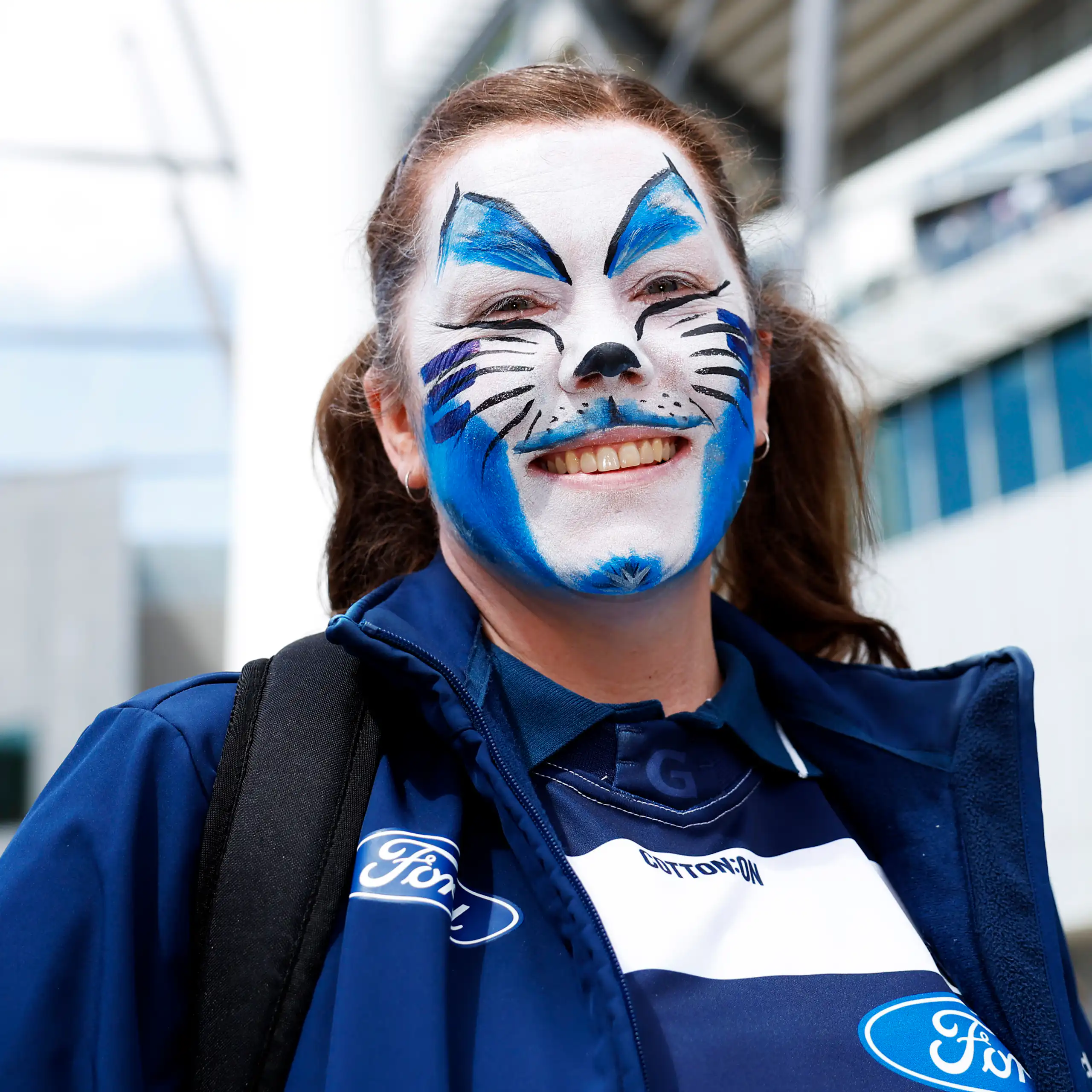 A young woman with blue and white cat face paint smiling outside a stadium.