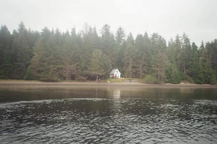 A small white hut surrounded by trees sits behind a large body of water.