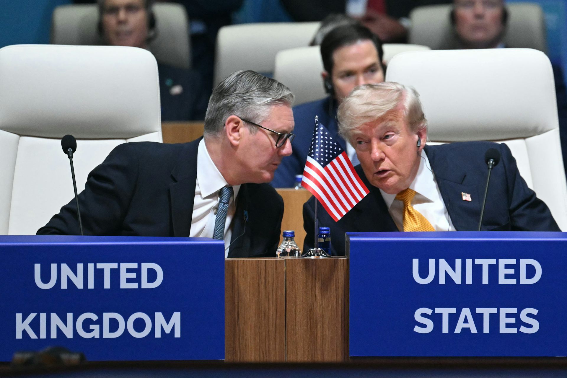 Two men in suits talk in front of signs that say United Kingdom and United States.