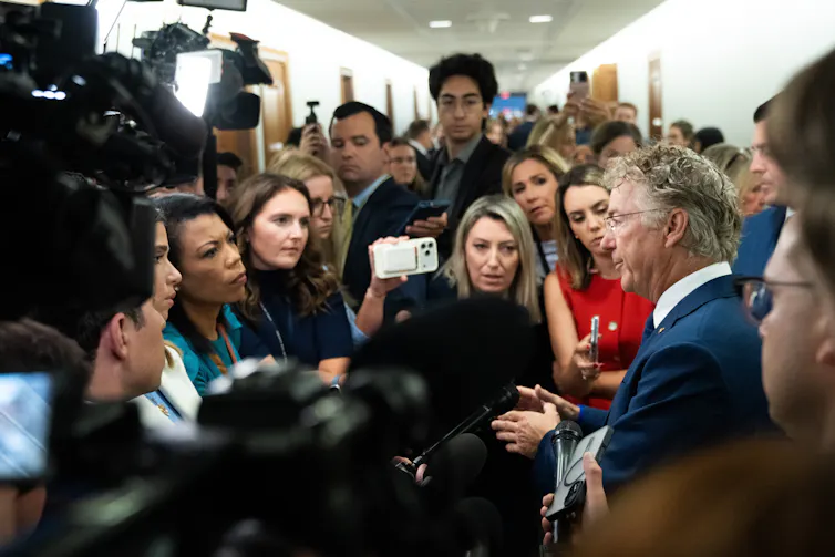 A man in a suit speaks to a crowd of reporters with phones, microphones and cameras in a hallway