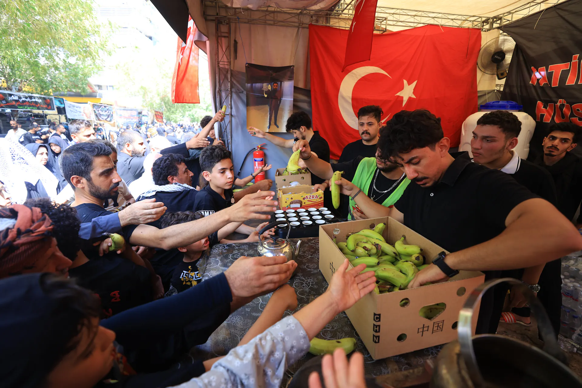 Several volunteers hand bananas from boxes to people reaching out to receive them. A Turkish flag with a white crescent and star is displayed on the side.