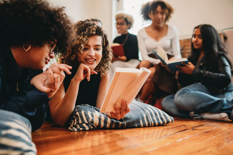 A group of young women either lie on floor pillows or sit on a sofa as they read and discuss books.