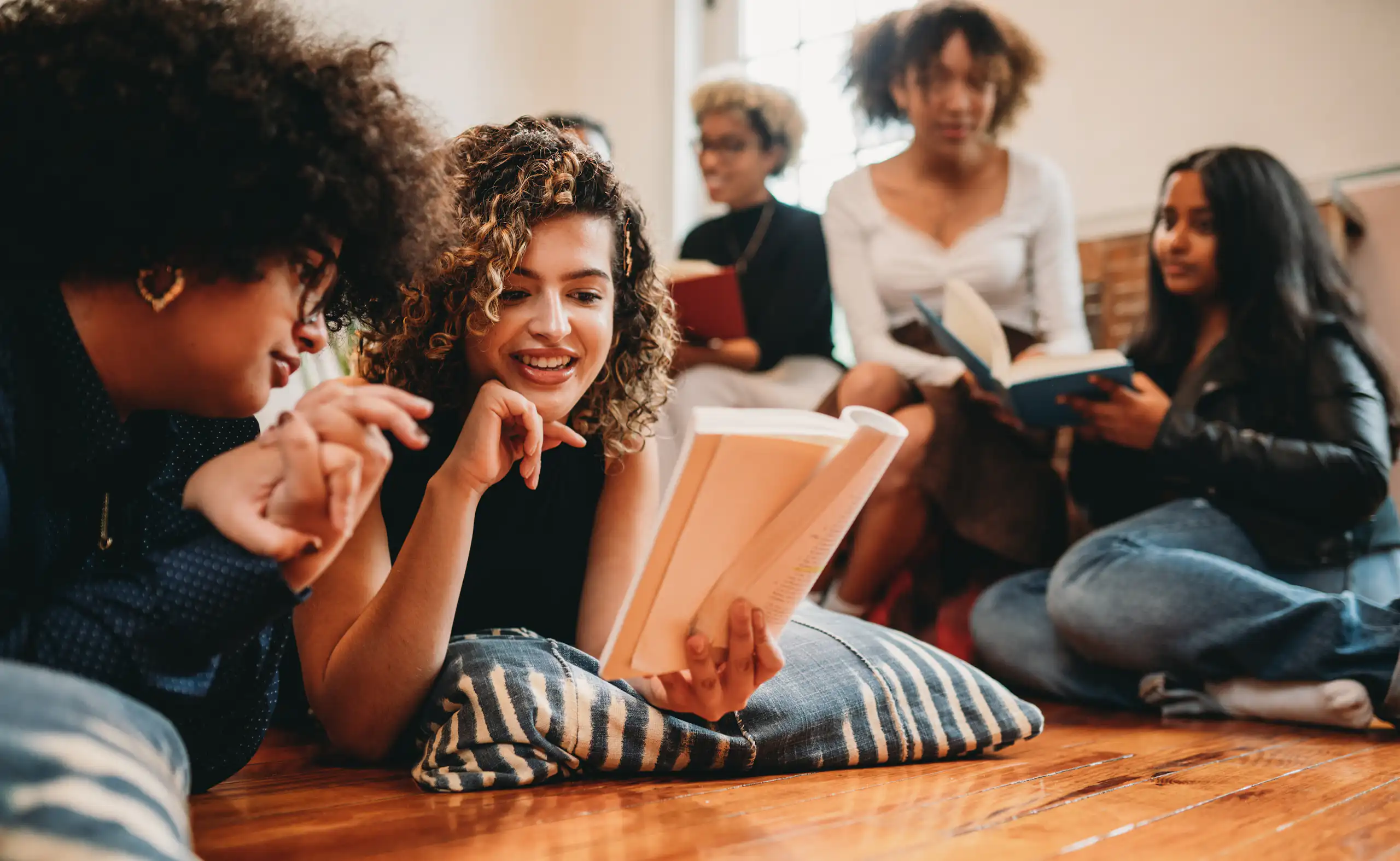 A group of young women either lie on floor pillows or sit on a sofa as they read and discuss books.