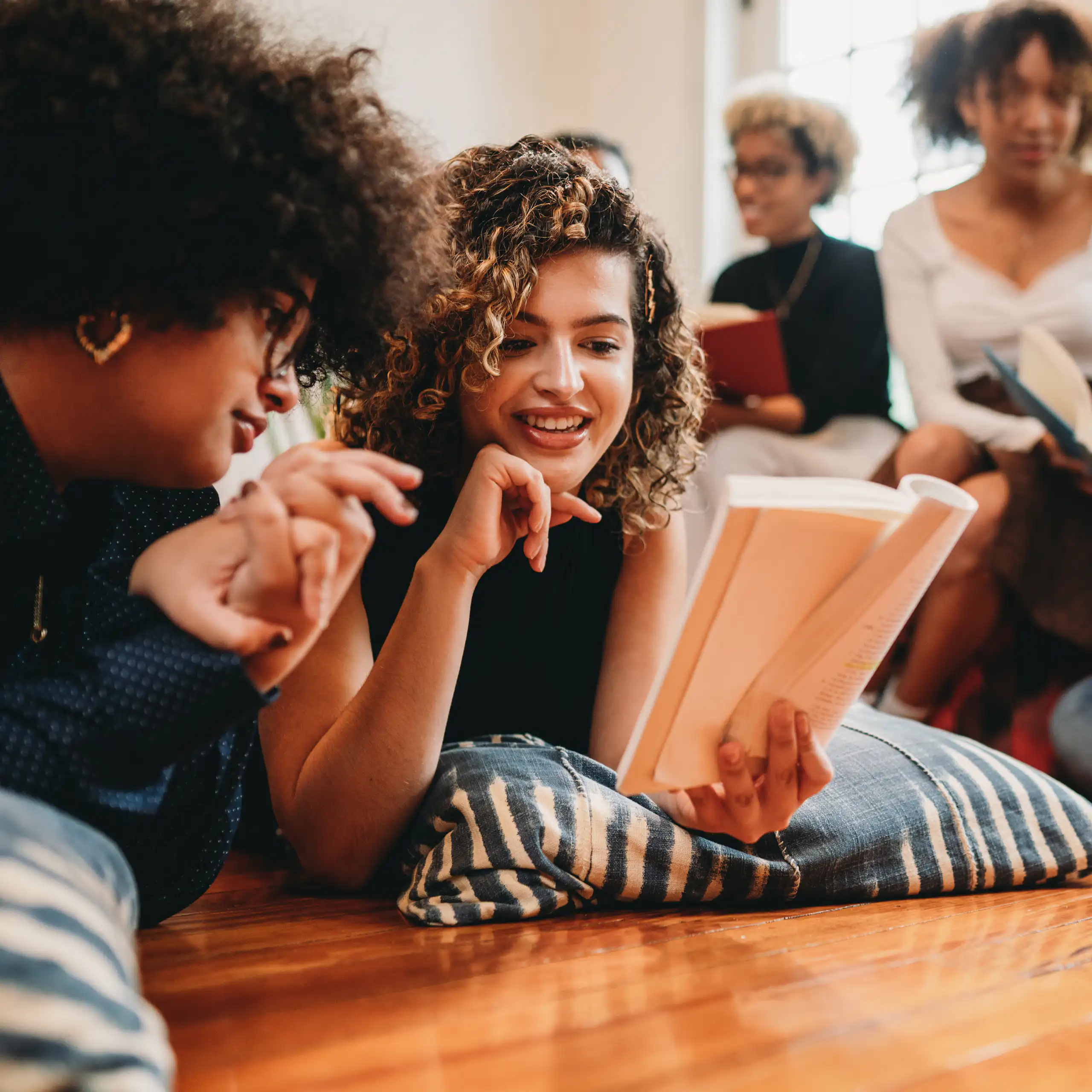A group of young women either lie on floor pillows or sit on a sofa as they read and discuss books.