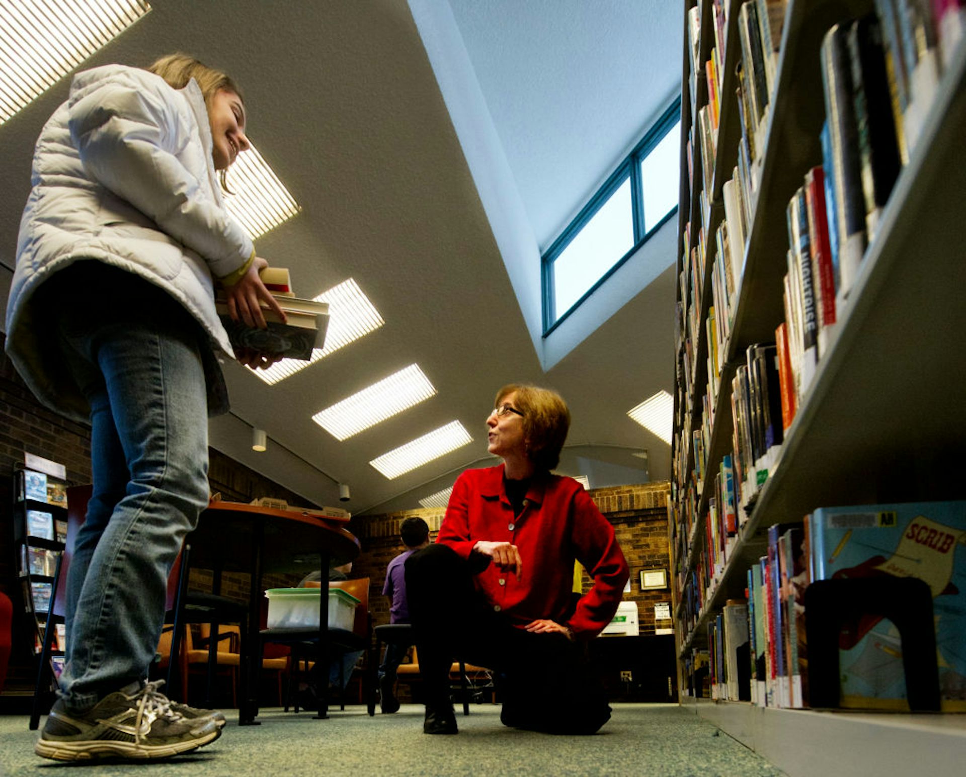 A teenage girl smiles while holding a stack of books and interacting with an older, female librarian who's crouching on one knee next to some bookshelves.