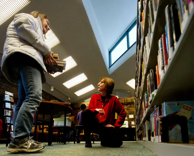 A teenage girl smiles while holding a stack of books and interacting with an older, female librarian who's crouching on one knee next to some bookshelves.