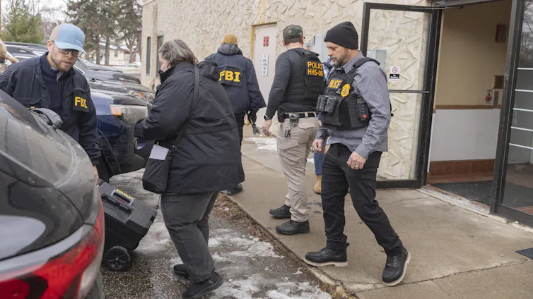 Agents, many wearing jackets or vests emblazoned with 'FBI,' exit a building with what appears to be a trove of documents.Medicaid Funding.