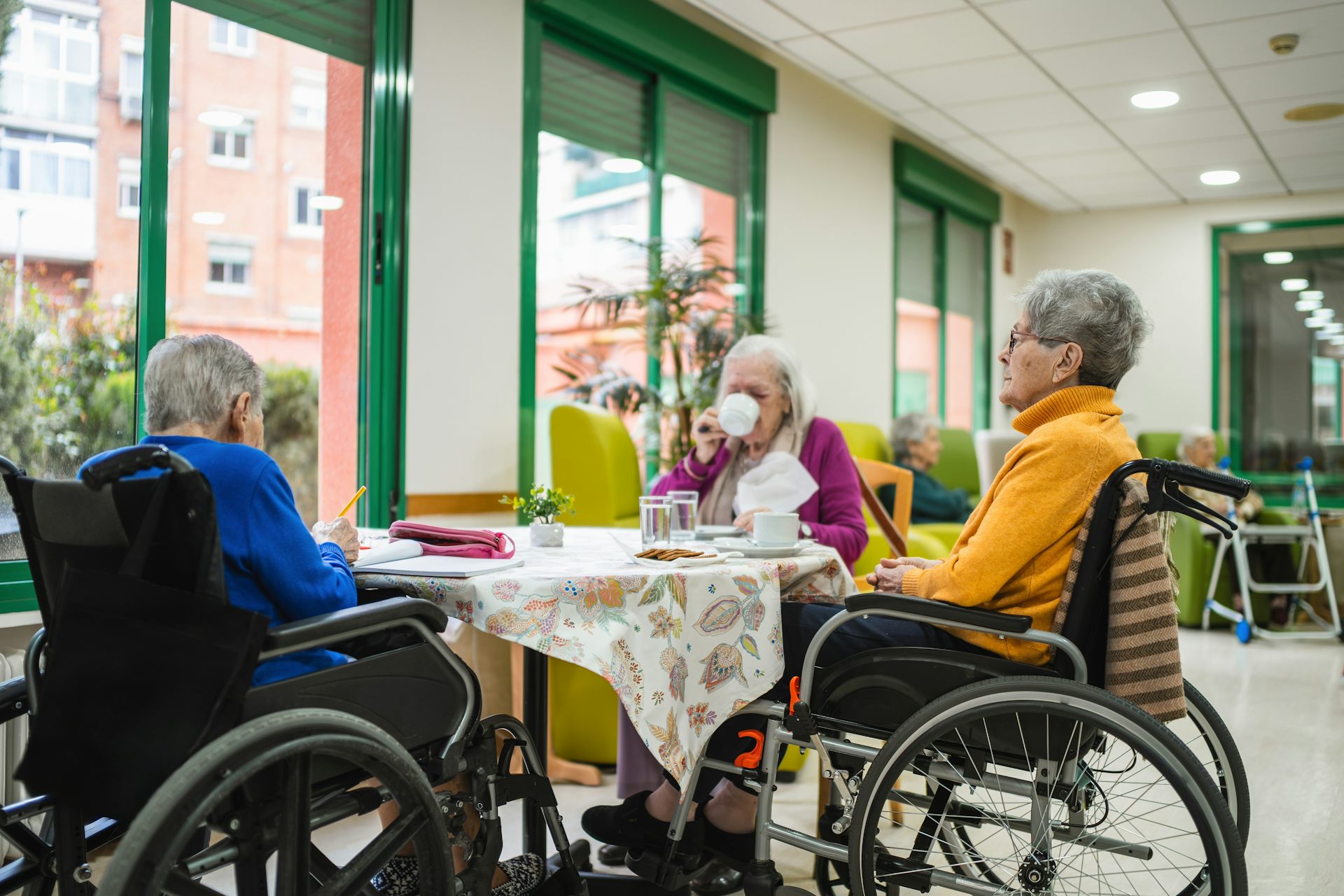 Three elderly women in wheelchairs sit around a table having tea.