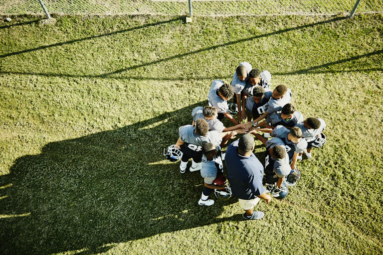 A group of young football players with blue jerseys and their coach stand in a circle and place their hands on top of one another in the middle.