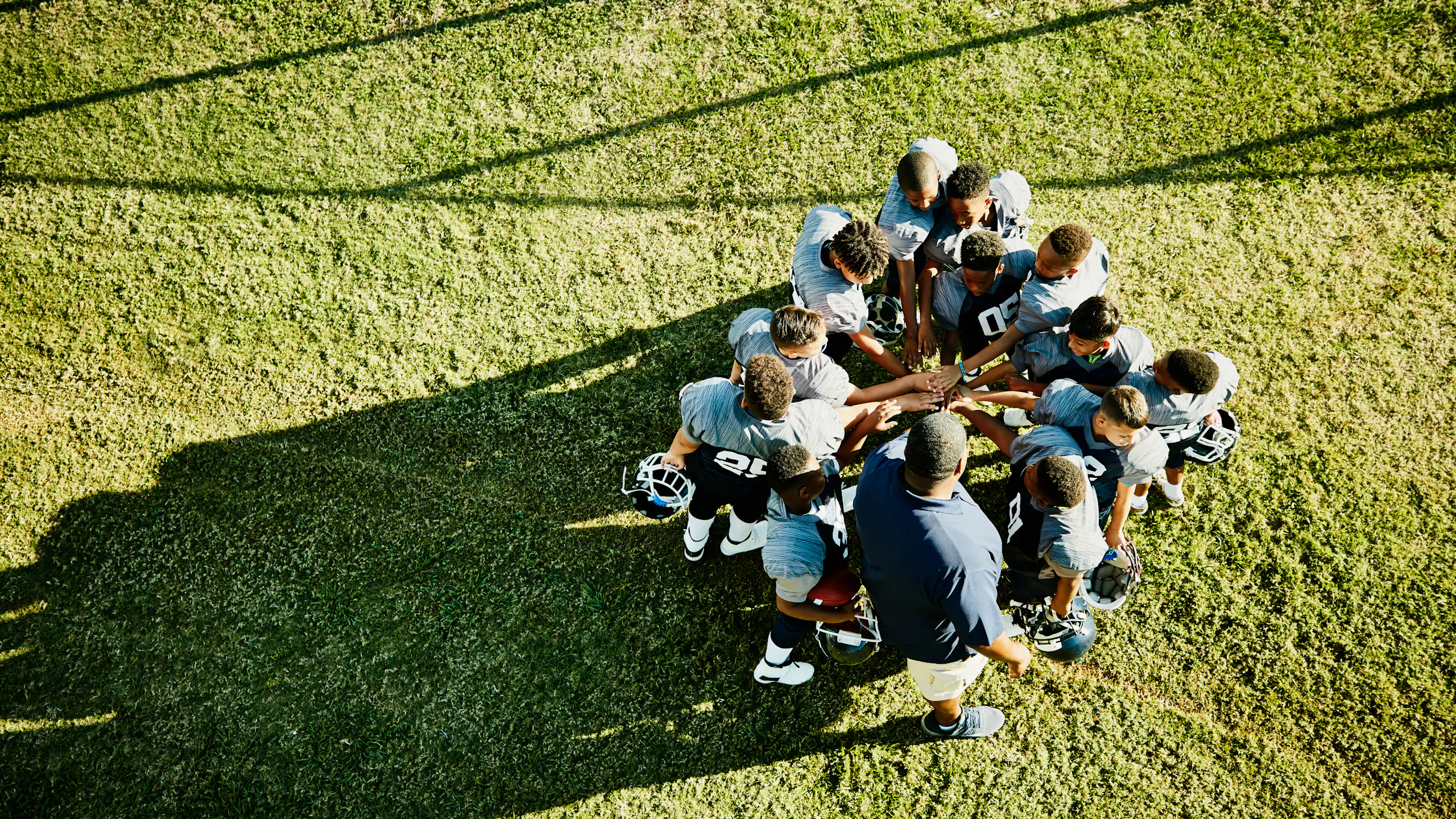 A group of young football players with blue jerseys and their coach stand in a circle and place their hands on top of one another in the middle.