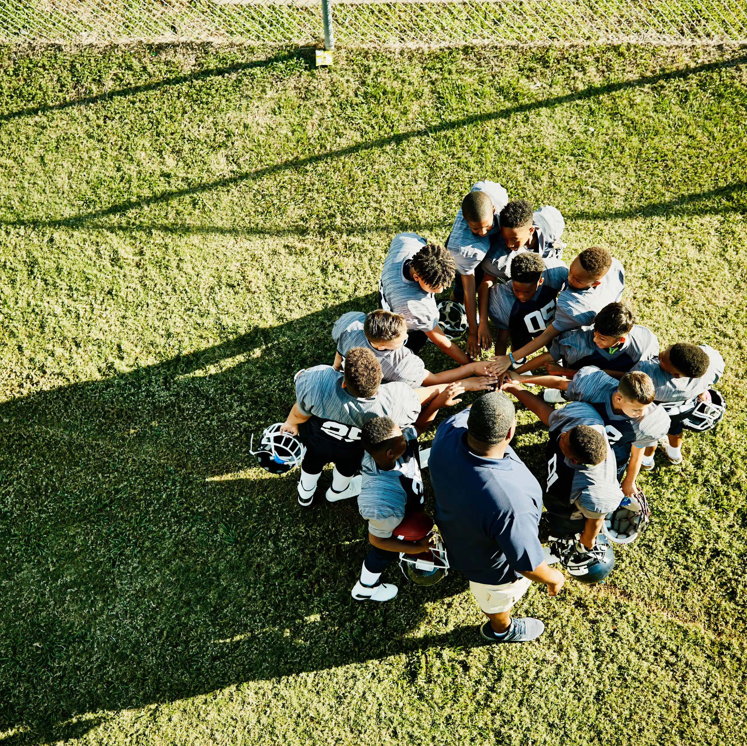 A group of young football players with blue jerseys and their coach stand in a circle and place their hands on top of one another in the middle.