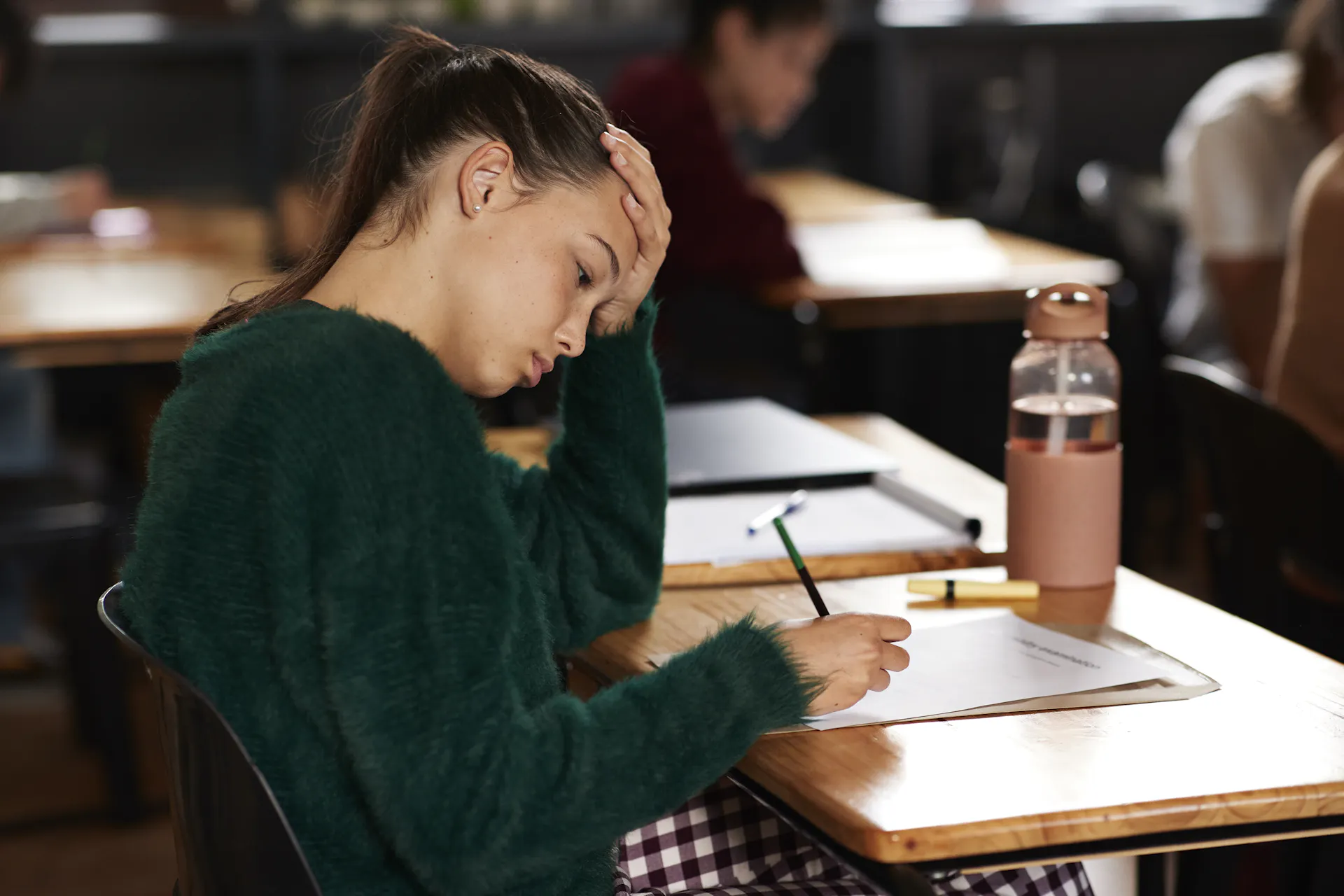 A teenage girl holds a pencil and leans one hand on her head while she sits at a desk. Other children are seen around her.