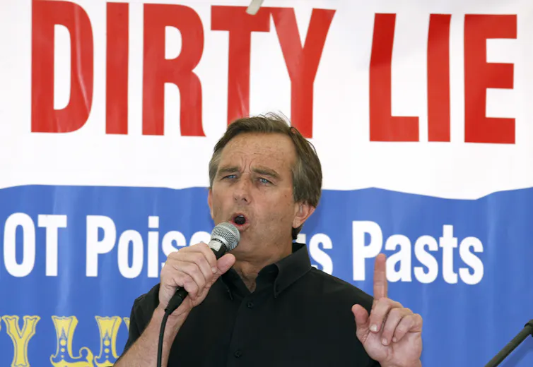 A man speaks into a microphone and gestures with his other hand in front of a sign saying 'dirty lie.'