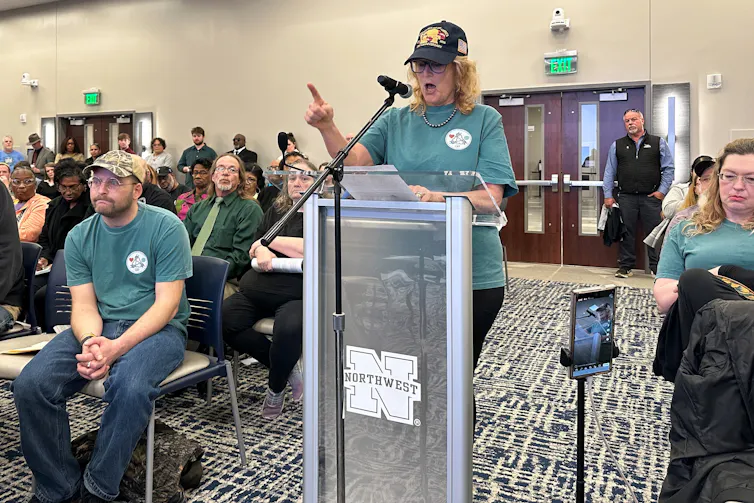 A person speaks at a lectern in a public meeting room.