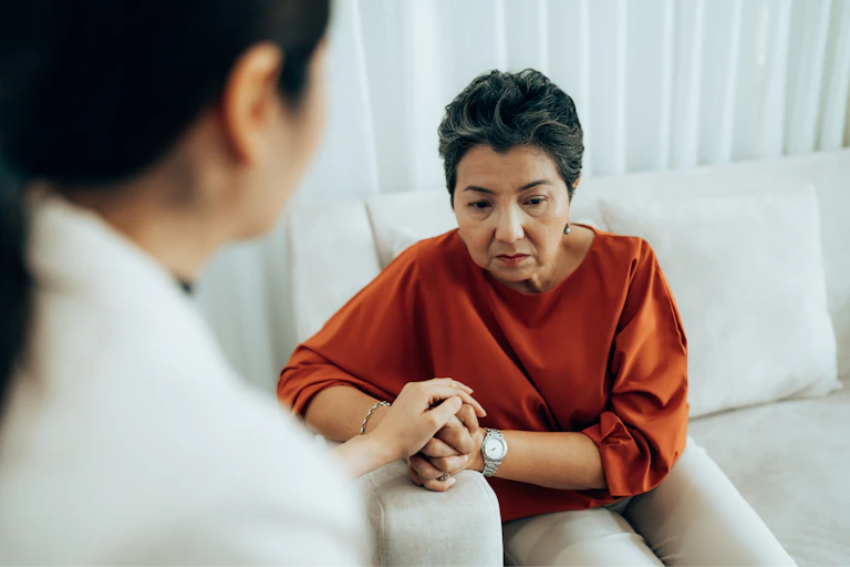 Senior Asian woman leaning forward on a couch during a therapy session, looking concerned.