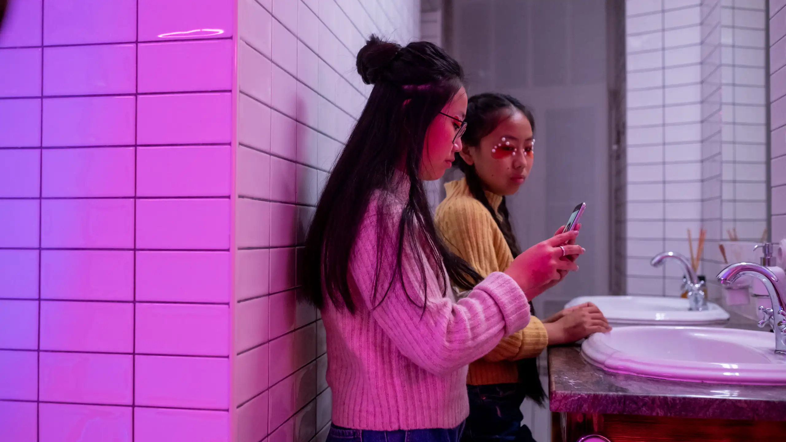 Two young women in a purple-lit bathroom using smartphones near a sink.