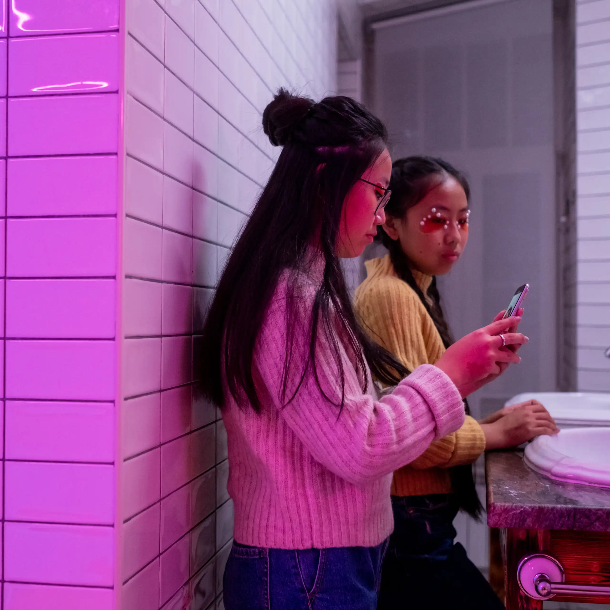 Two young women in a purple-lit bathroom using smartphones near a sink.