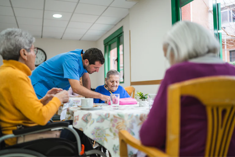A male care worker wearing blue scrubs speaks to an elderly care resident alongside two other elderly women who are sitting around the same table.