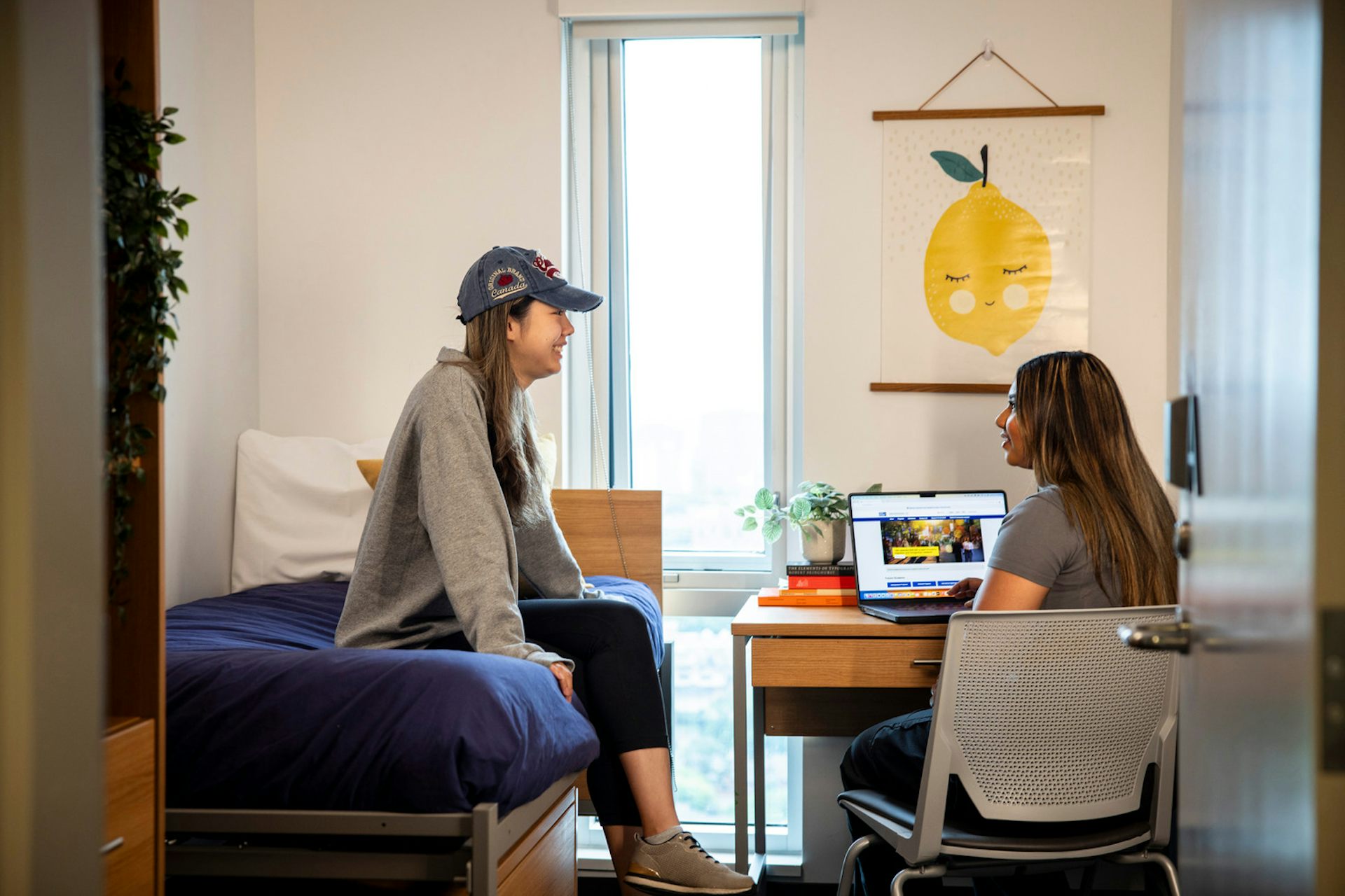 Two students talk to each other in a dorm room. One works on a laptop and other sits on bed.