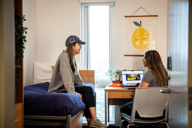 Two students talk to each other in a dorm room. One works on a laptop and other sits on bed.